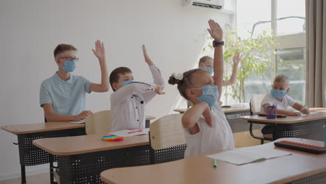 children-in-the-classroom-at-school-in-masks-sit-in-the-classroom-and-answer-the-teacher's-questions-by-raising-their-hands-in-slow-motion.-Lessons-during-the-pandemic-at-school